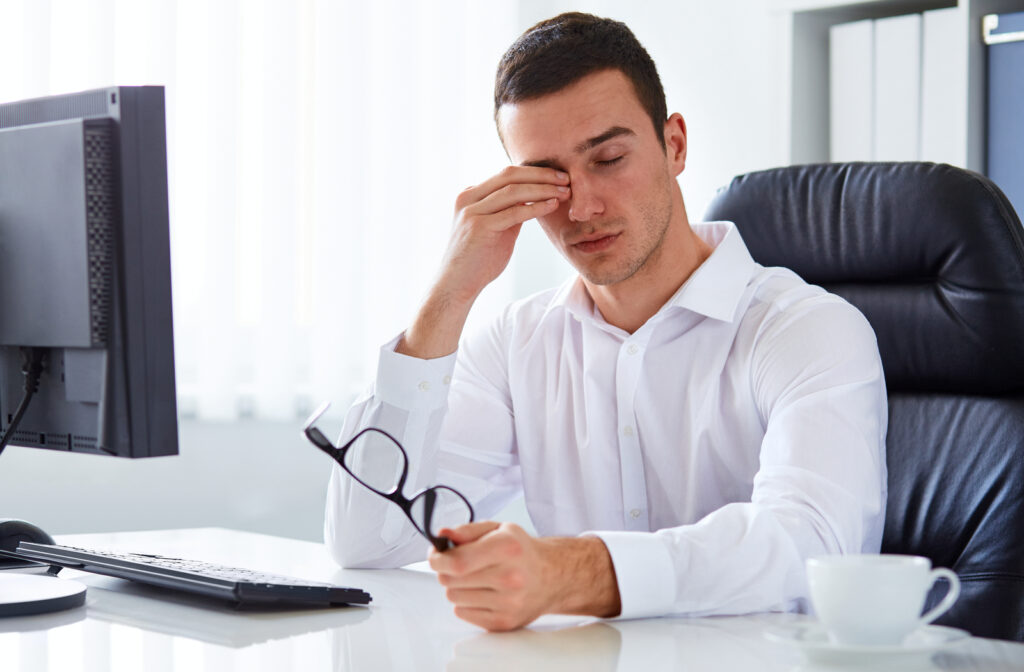 Young man experiencing tired eye while working with computer at work desk while taking off glasses