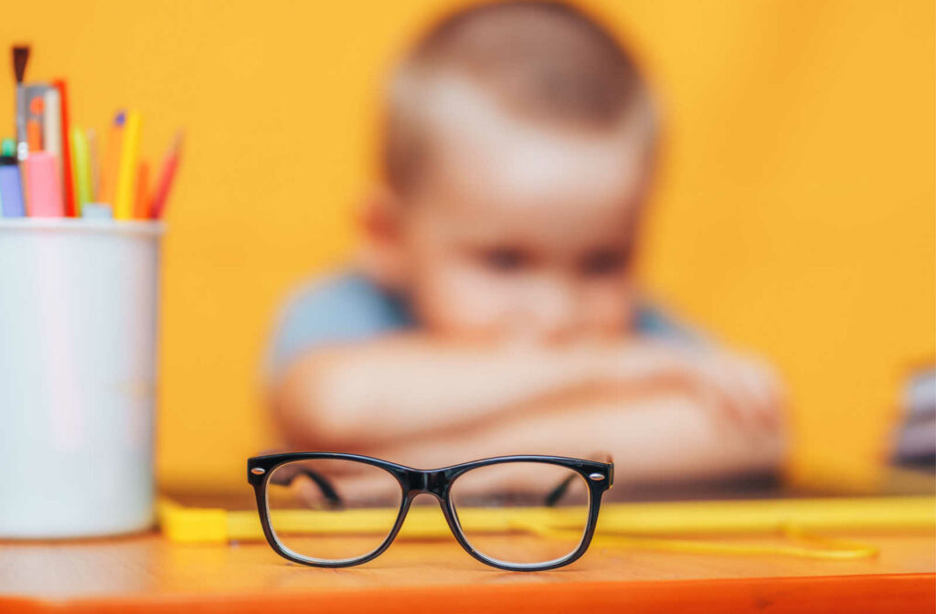 A young boy blurred in the background with his head rested on his crossed arms struggles to see clearly in class due to myopia while glasses sit on the desk in focus.