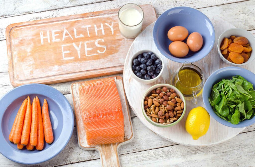 A variety of eye-healthy foods, including salmon, carrots, eggs, milk, and nuts, arranged on platters with a cutting board that has "healthy eyes" written in flour at the top.
