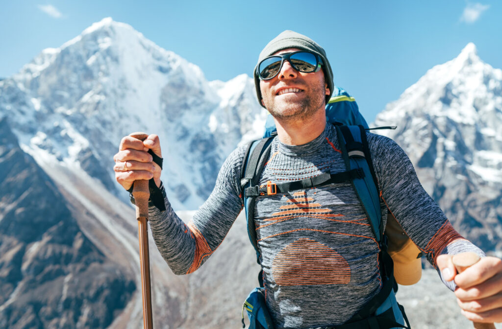 A man on top of a mountain wearing Polarized sunglasses and a backpack while hiking with poles.