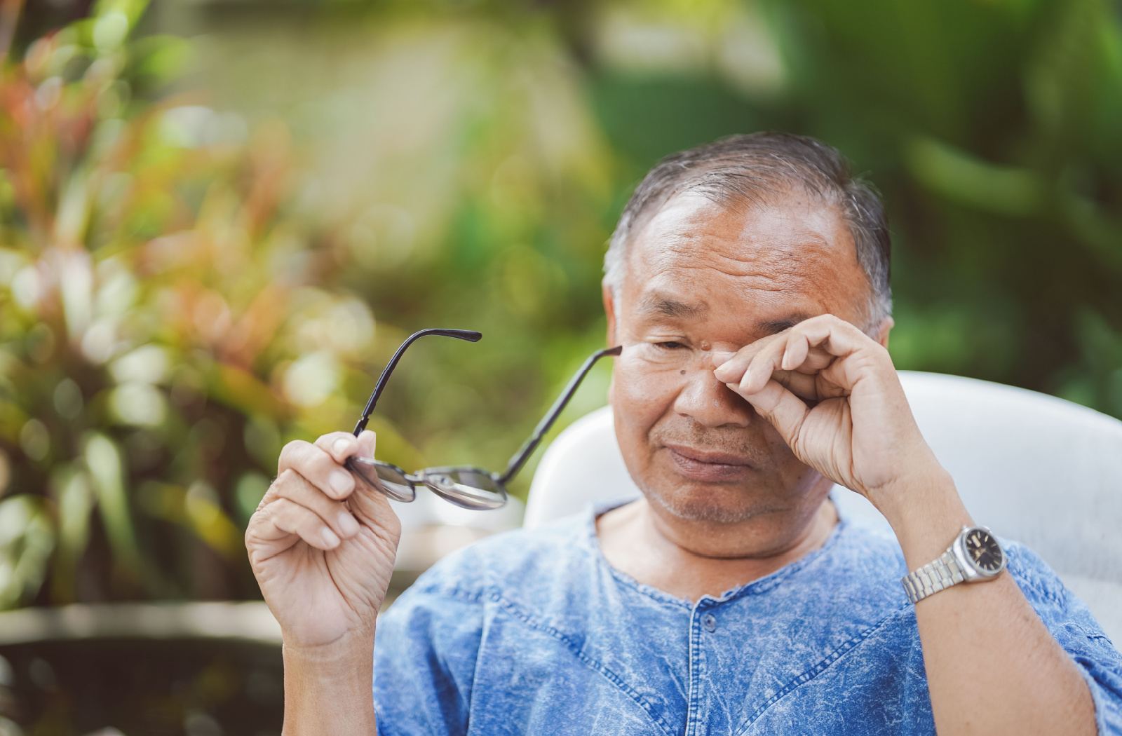 A senior man sitting outside in a chair holding his glasses in his right hand as he rubs his left eye with his left hand