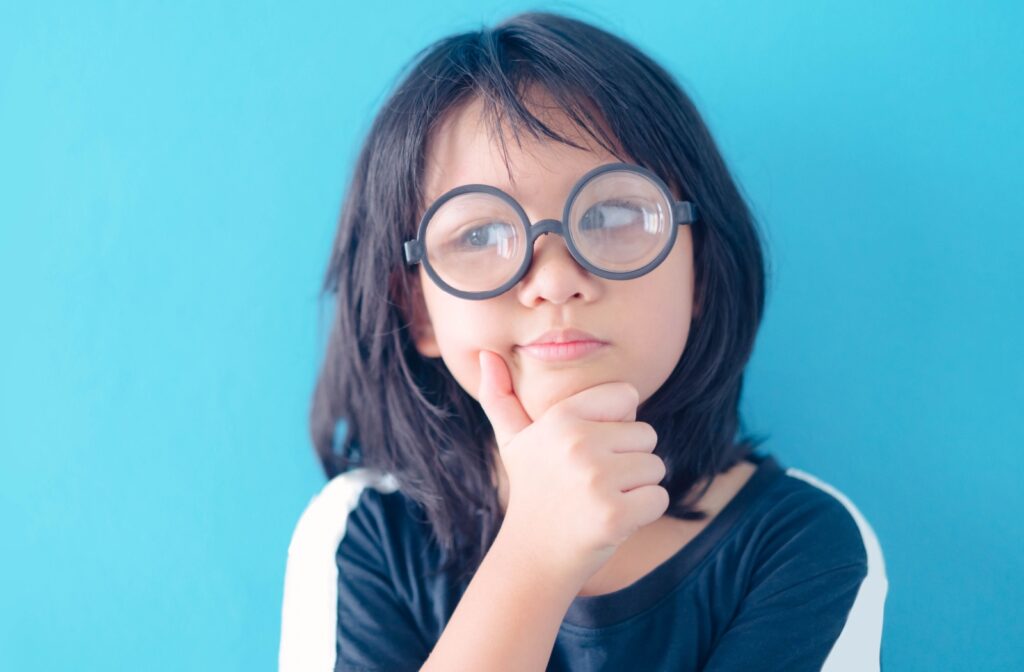 A young child with black hair and glasses has their hand on their chin thinking.