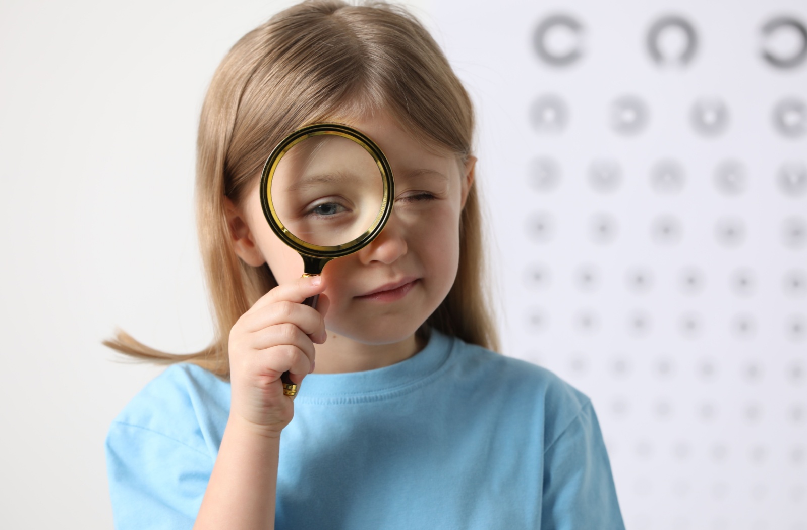 A young child wearing a blue shirt holds a magnifying glass in front of their eyes.