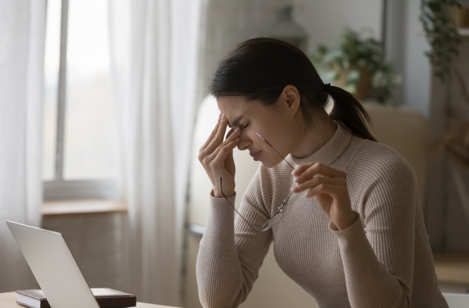 A person sitting in front of a laptop, holding their glasses in 1 hand and rubbing their eyes due to dry eyes