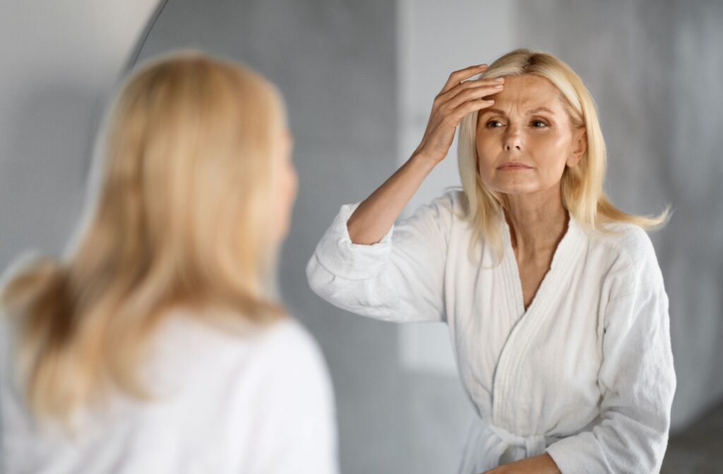 A person in a white dressing gown looking in the mirror and examining their face for wrinkles and dark spots