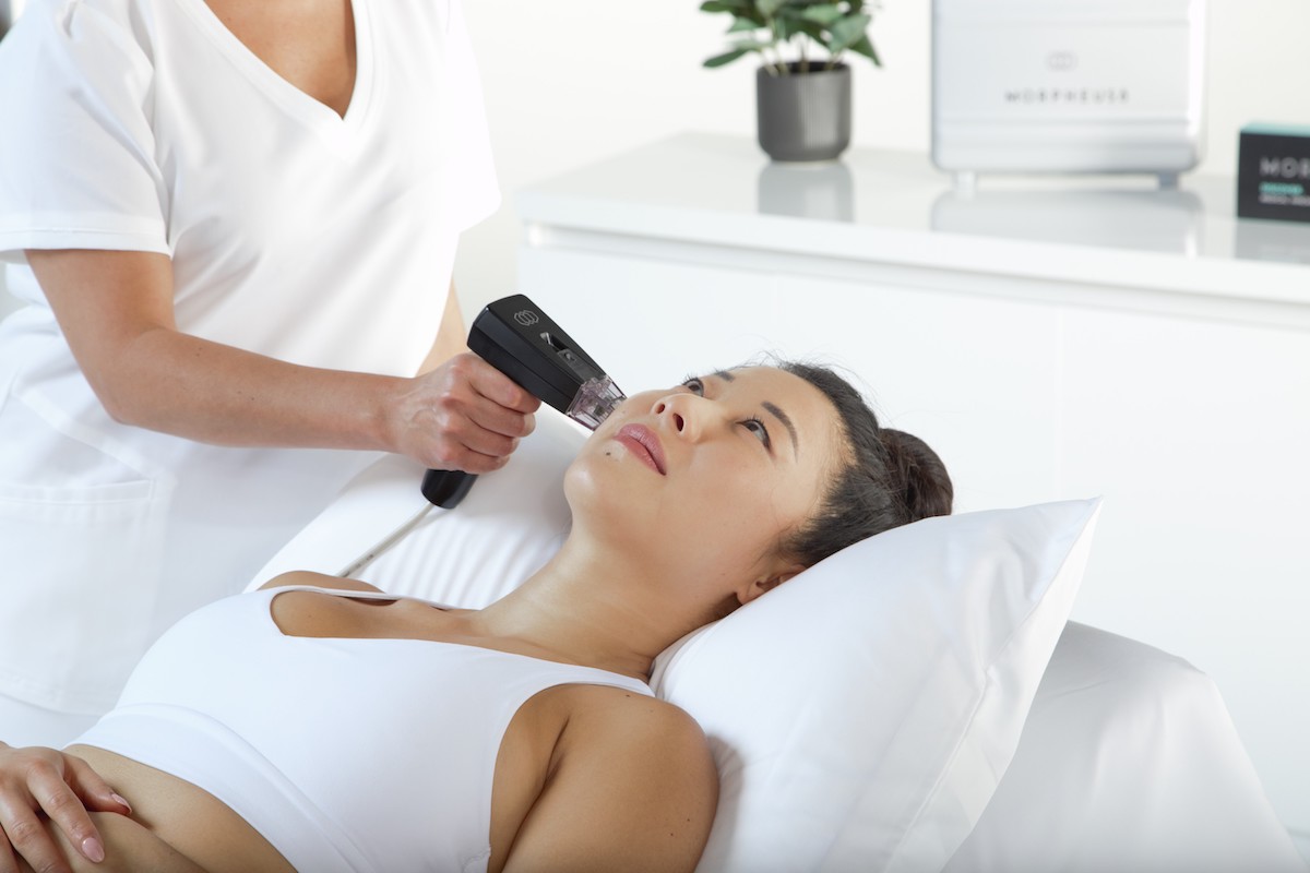 Clinician using a handheld aesthetic treatment device on a woman’s cheek as she lies on a treatment bed in a bright clinic.