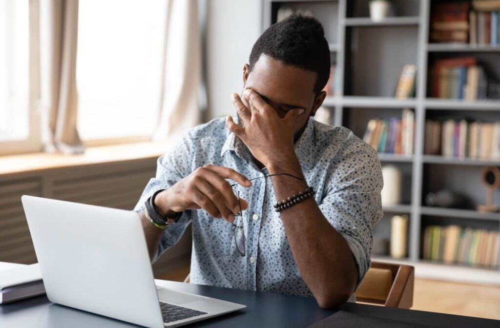 A person holding their glasses in 1 hand and their eyes with their other hand due to strained eyes while working on a laptop.