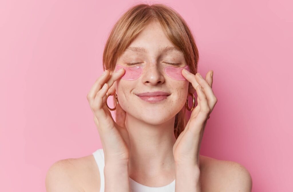 Close-up of a person against a pink background smiling with their eyes closed and pressing on pink under-eye patches.