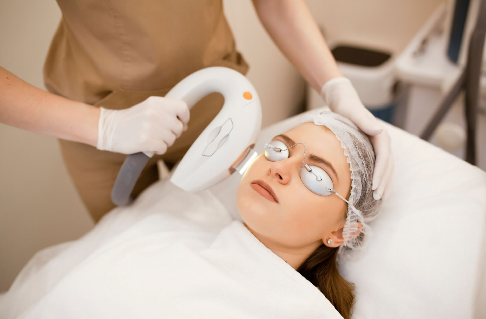 A female patient is lying in an operating bed with her eyes covered by laser-protective goggles and a hair net on her head whit the female doctor is performing an IPL on her face to relieve the patient's dry eyes.