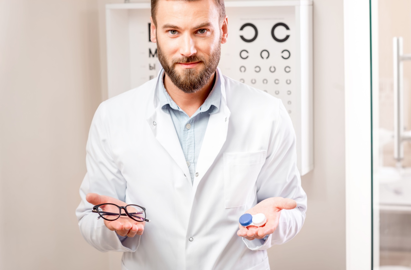 An optometrist holds eyeglasses in one hand and a contact lens in the other.