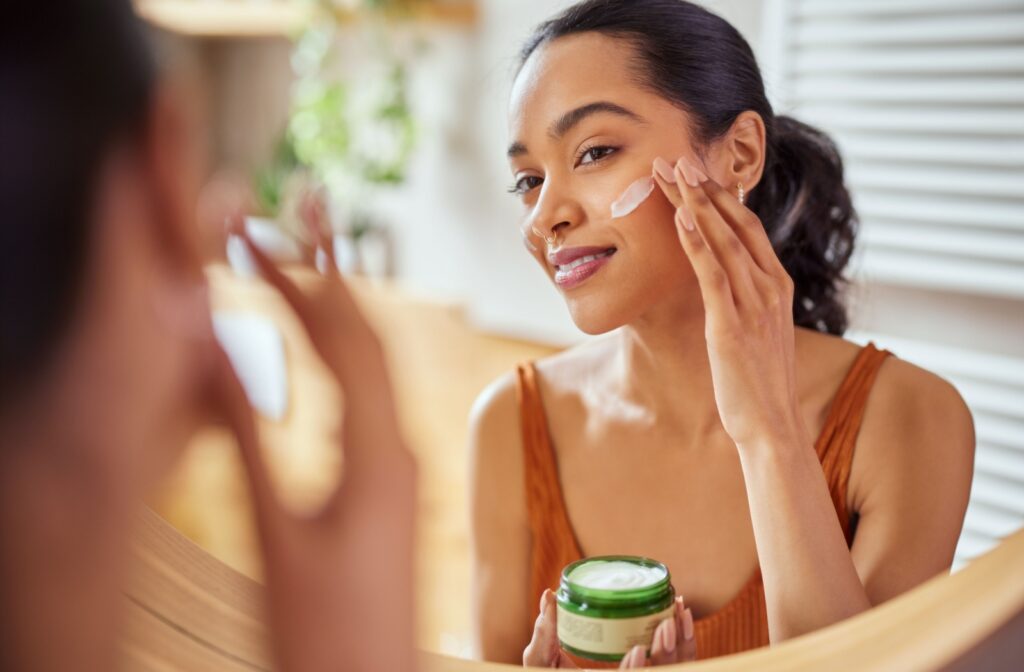A smiling adult puts a high quality cream on their face as part of their skincare routine.