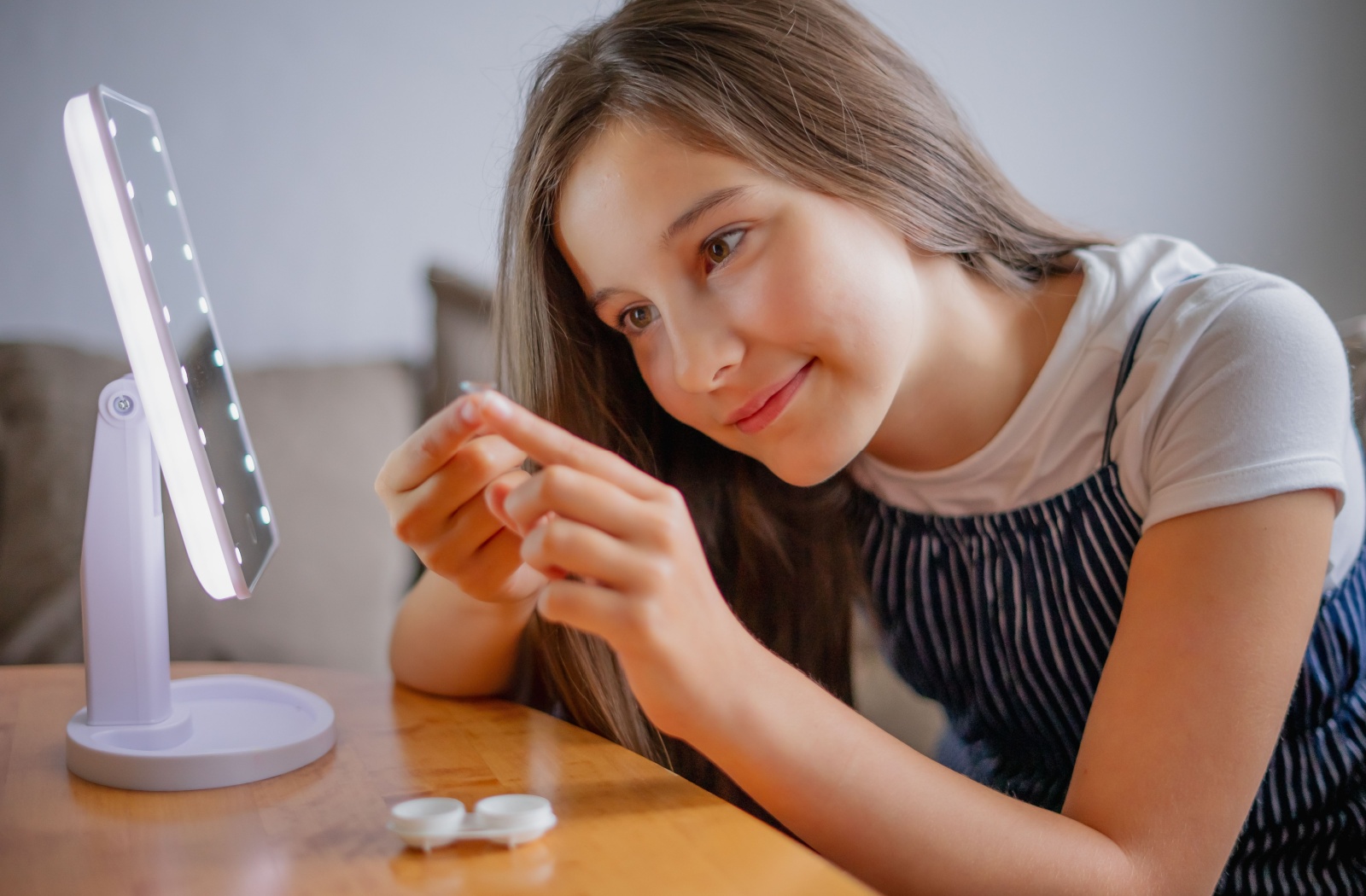 Smiling girl sitting at a table with a mirror, holding a contact lens on her finger while practicing insertion.
