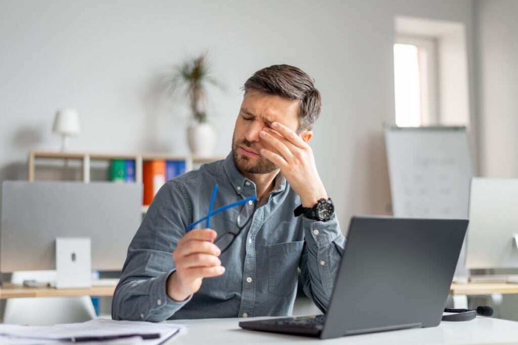Professional man at desk rubbing his eye and temple while working on laptop, demonstrating eye fatigue, headache, and dry eye symptoms from prolonged screen time and digital eye strain.