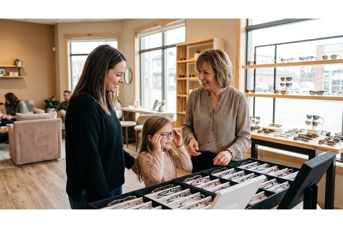 A friendly optical consultant helping a young girl try on eyeglasses while her mother looks on in a bright, modern optometry office, with racks of eyewear visible in the background.